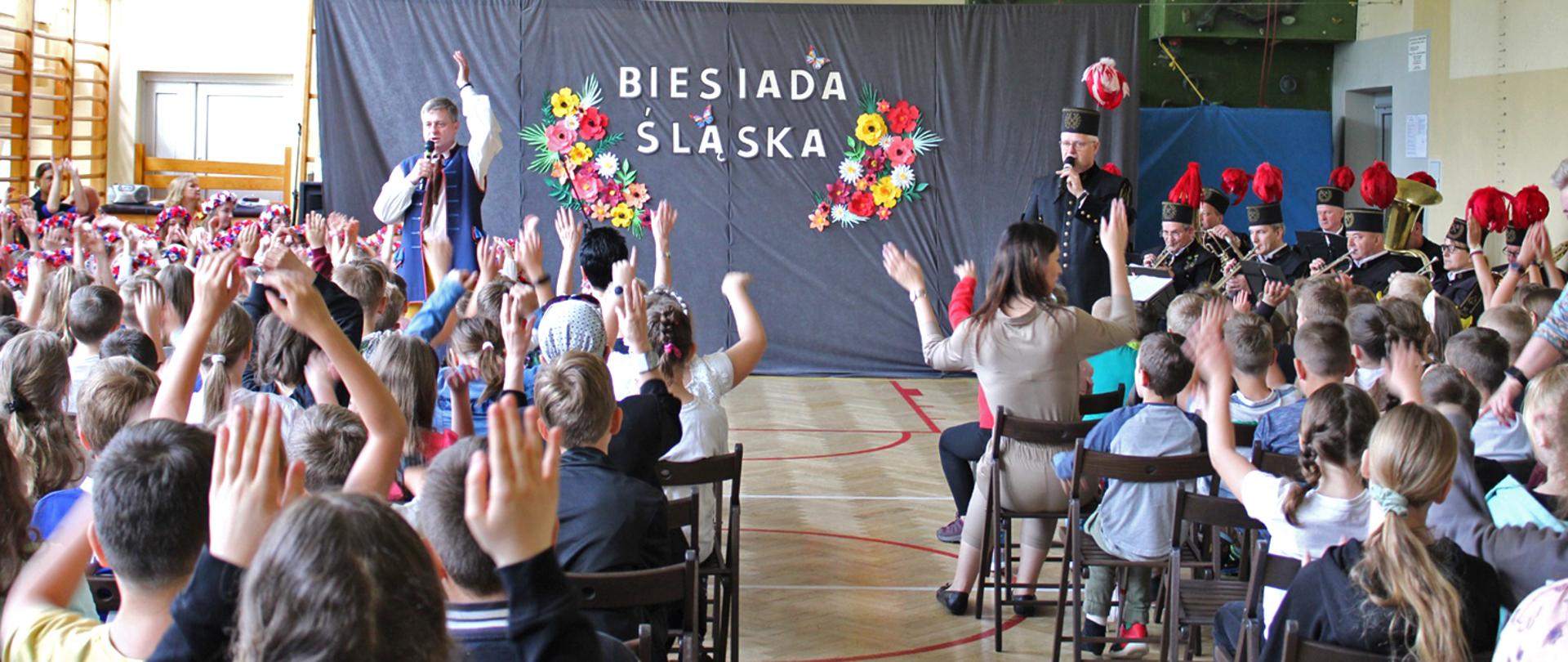 Grupa uczniów biorąca udział w biesiadzie na sali gimnastycznej. W tle prowadzący biesiadę oraz orkiestra górnicza a za nimi biały napis biesiada śląska obok napisu kompozycje kwiatowe.