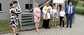 Przed budynkiem szkoły stoją od lewej: nauczycielka prowadząca uroczystość, pani dyrektor szkoły, ksiądz poświęcający nowe logo szkoły, dwóch ministrantów i wicewójt gminy Łabowa