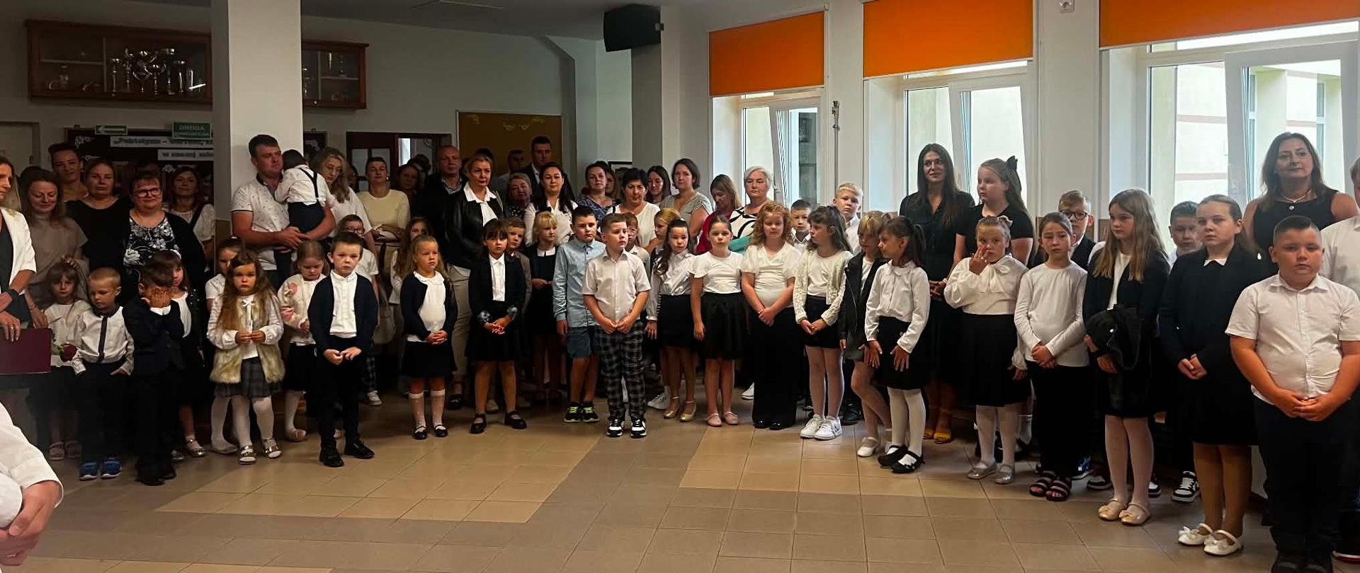 Uczniowie wraz z nauczycielami i rodzicami stoją na korytarzu szkolnym. Dzieci ubrane są w stroje galowe, zaś dorośli - odświętnie. 