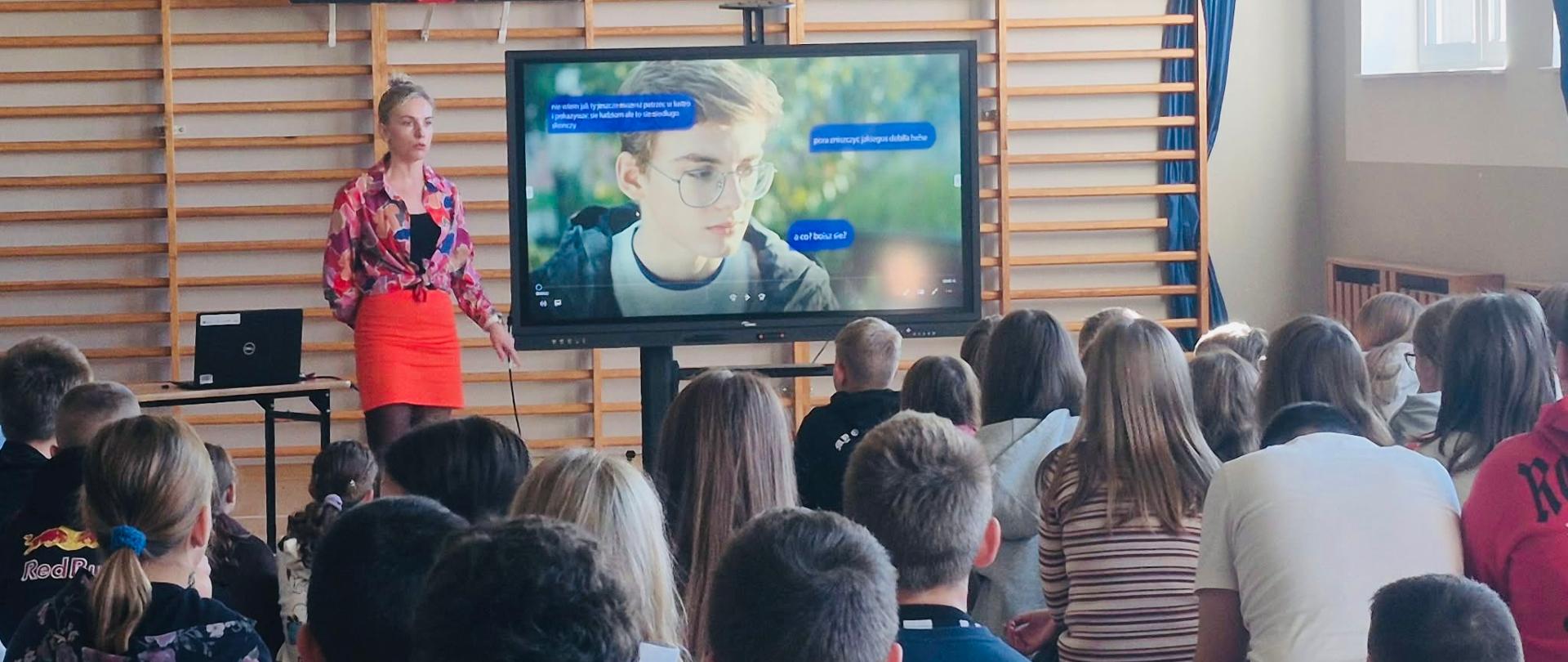 Prowadząca zajęcia w kolorowej koszuli i pomarańczowej spódnicy stoi przy ekranie w sali gimnastycznej. Na ekranie widać kadr z filmu: chłopca w okularach oraz wiadomości czatu, w których ktoś go wyśmiewa. Uczniowie siedzą przodem do ekranu.