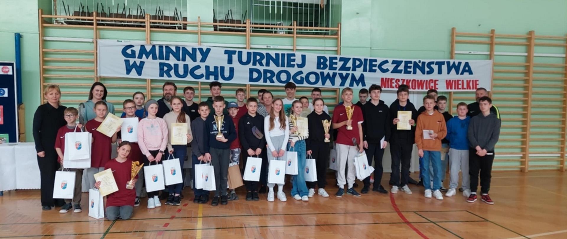 W szkolnej sali gimnastycznej przy drabinkach do zdjęcia pozuje duża grupa uczniów wraz z nauczycielami opiekunami. Uczniowie trzymają w rękach nagrody, dyplomy i puchary. Nad nimi widnieje podłużny baner z napisem Gminny Turniej Bezpieczeństwa w Ruchu Drogowym.