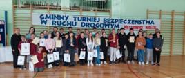 W szkolnej sali gimnastycznej przy drabinkach do zdjęcia pozuje duża grupa uczniów wraz z nauczycielami opiekunami. Uczniowie trzymają w rękach nagrody, dyplomy i puchary. Nad nimi widnieje podłużny baner z napisem Gminny Turniej Bezpieczeństwa w Ruchu Drogowym.