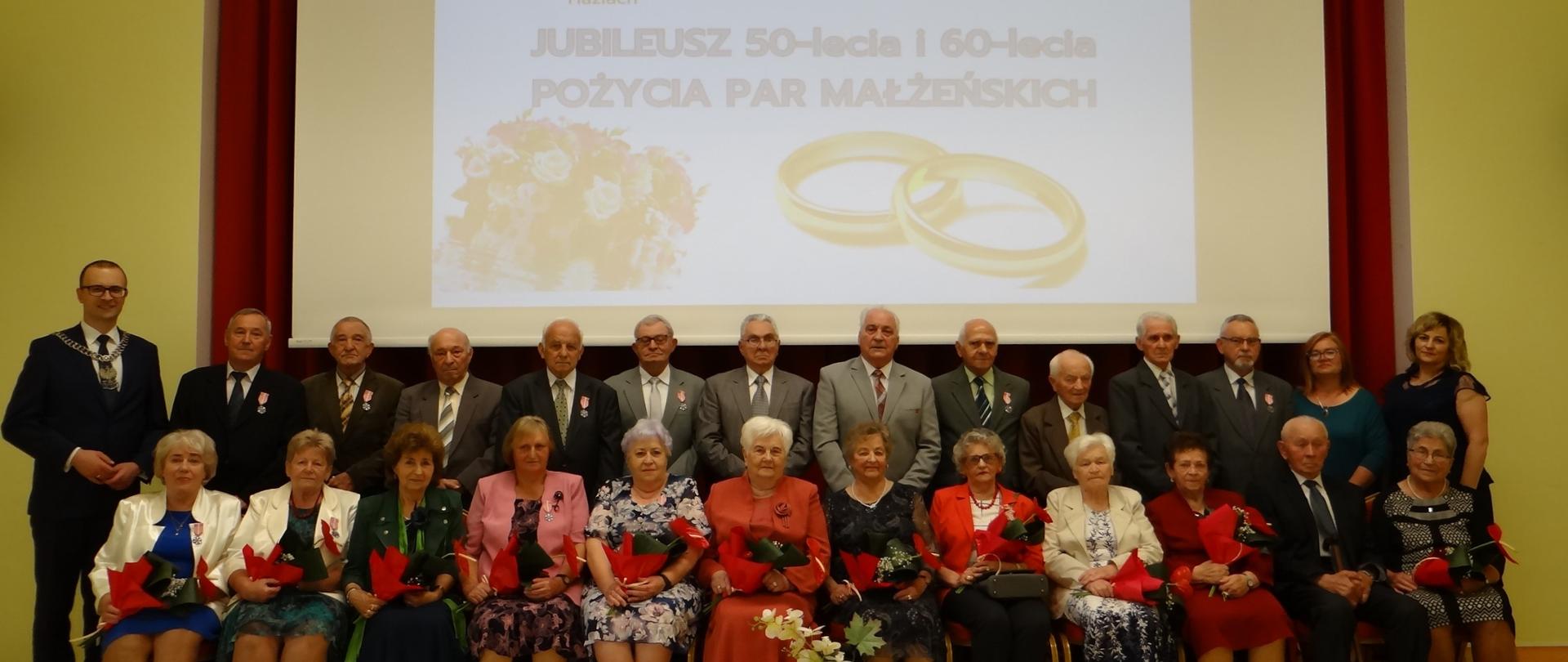 Zacni Jubilaci 50 i 60 lat pożycia małżeńskiego. Zbiorowa fotografia wykonana w sali widowiskowej Gminnej Biblioteki Publicznej. Osoby w pierwszym rzędzie siedzą na krzesłach, drugi rząd stoi.