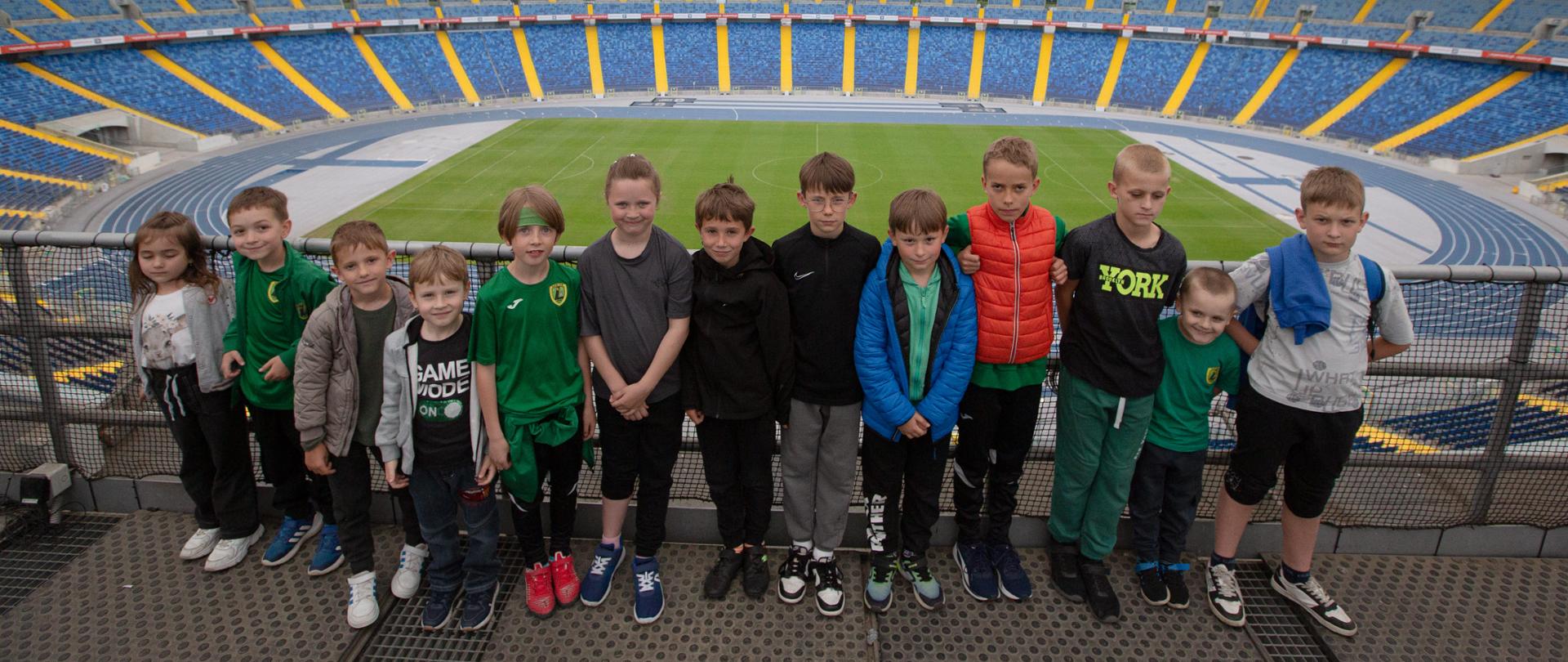 Dzieci pozują do zdjęcia na stadionie w tle zielona murawa i niebieskie krzesełka widowni