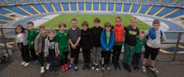 Dzieci pozują do zdjęcia na stadionie w tle zielona murawa i niebieskie krzesełka widowni