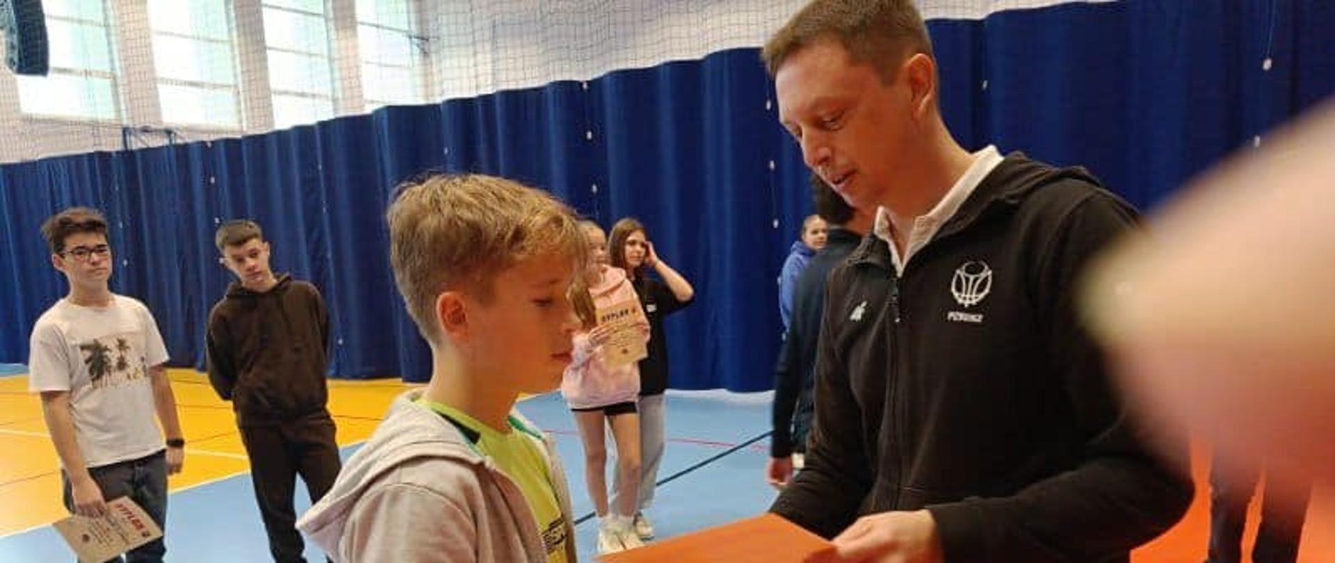 Na hali Marcin w szarej dresowej bluzie odbiera z rąk pana P. Lizaka ubranego w ciemny dres dyplom.