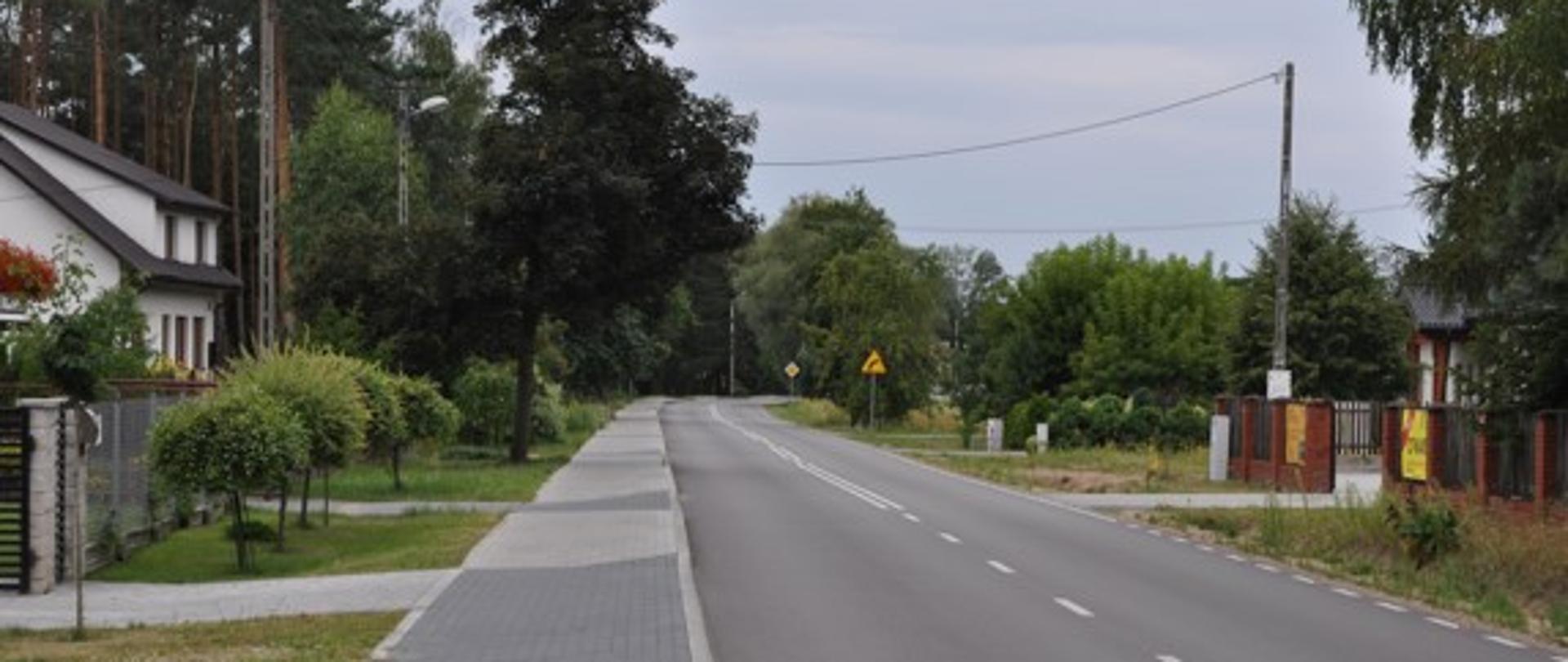 Na zdjęciu widnieje fragment wyremontowanej drogi powiatowej wraz z chodnikiem. Po obu stronach drogi znajdują się zabudowania oraz roślinność.