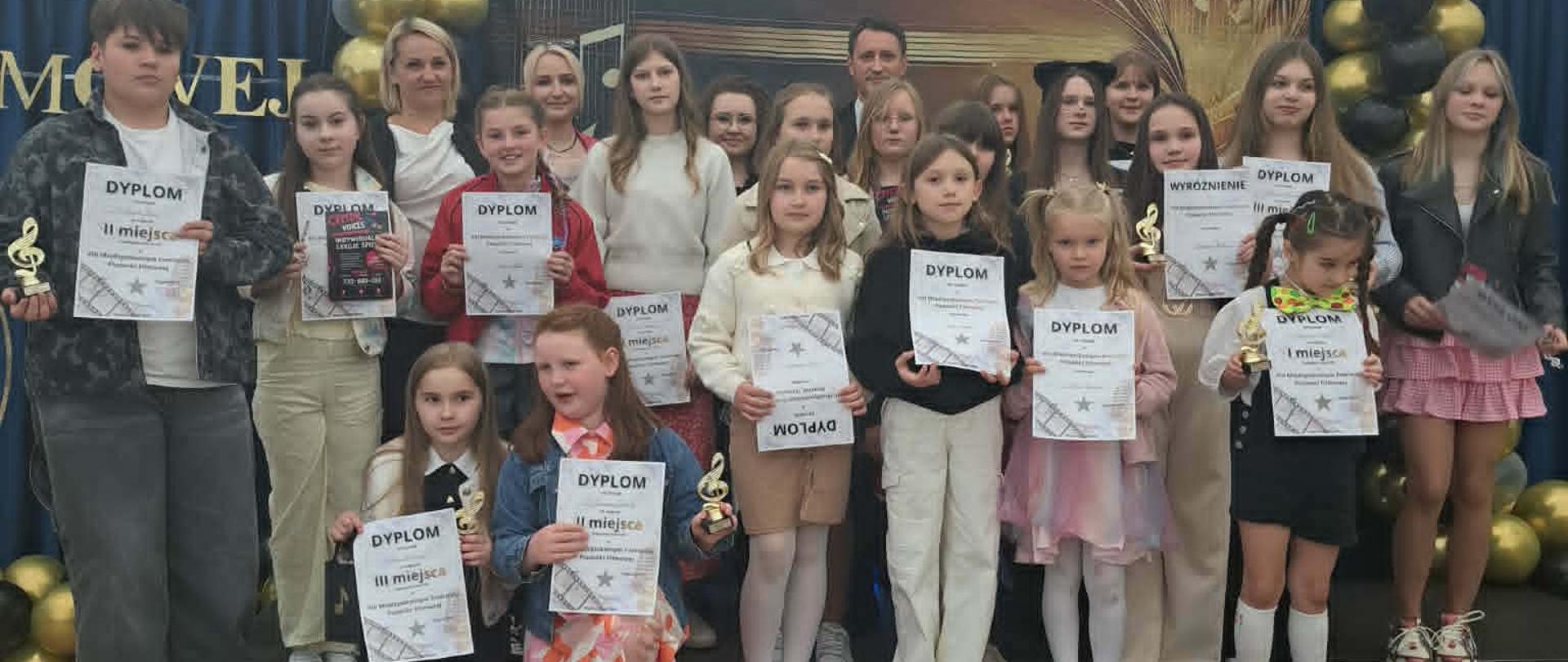 Grupa dzieci i dorosłych stoi na scenie, pozując do wspólnego zdjęcia po zakończeniu festiwalu muzycznego. Uczestnicy trzymają w dłoniach pamiątkowe dyplomy oraz małe statuetki w kształcie klucza wiolinowego. W tle widoczna jest elegancka dekoracja z czarnych i złotych balonów oraz fragment napisu na ciemnoniebieskim materiale.