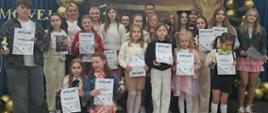 Grupa dzieci i dorosłych stoi na scenie, pozując do wspólnego zdjęcia po zakończeniu festiwalu muzycznego. Uczestnicy trzymają w dłoniach pamiątkowe dyplomy oraz małe statuetki w kształcie klucza wiolinowego. W tle widoczna jest elegancka dekoracja z czarnych i złotych balonów oraz fragment napisu na ciemnoniebieskim materiale.