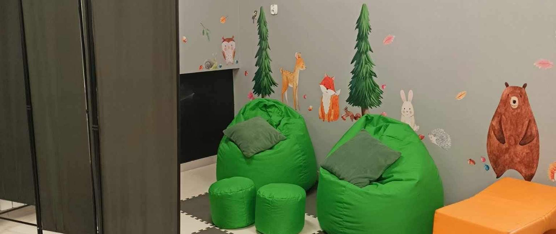 Na podłodze maty biało-szare, na nich stoją 2 zielone miękkie fotele, przed nimi 2 zielone miękkie pufy, po lewej stronie czarna tablica do pisania kredą, ściany szare, na nich naklejone wiewiórki, zielone świerki, ślimak, sowa, sarna, lisek, zając, sikorka i gil