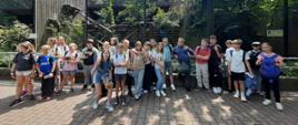 Grupa dzieci stojących przed dużą klatką w ogrodzie zoologicznym. Dzieci stoją w dwóch rzędach, niektórzy trzymają napoje lub przekąski. W tle widać gęstą roślinność, drewniane konstrukcje i siatkę zabezpieczającą.