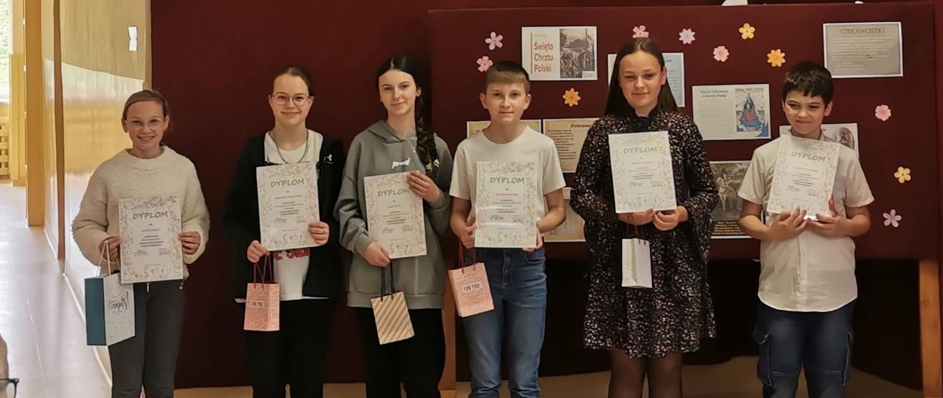 4 uczennice i 2 uczniów z klas od 4 do 8 stoi na tle bordowej dekoracji, trzymają w rękach dyplomy za Polsko-Angielski Konkurs Ortograficzny oraz nagrody w papierowych torebeczkach