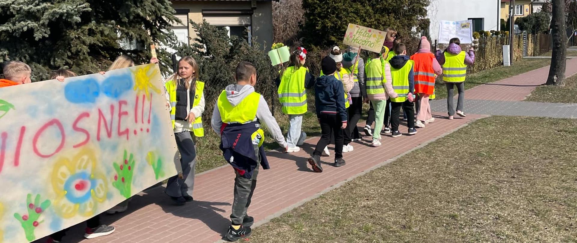 Grupa dzieci idąca chodnikiem. Część z nich ma założone zielone kamizelki odblaskowe. Widać niesiony baner z napisem wiosnę, kolorowymi kwiatami, słońcem i chmurami