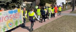 Grupa dzieci idąca chodnikiem. Część z nich ma założone zielone kamizelki odblaskowe. Widać niesiony baner z napisem wiosnę, kolorowymi kwiatami, słońcem i chmurami