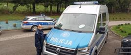 Przed budynkiem szkolnym stoi furgonetka policyjna i policyjny samochód osobowy, obok furgonetki stoi pani policjantka