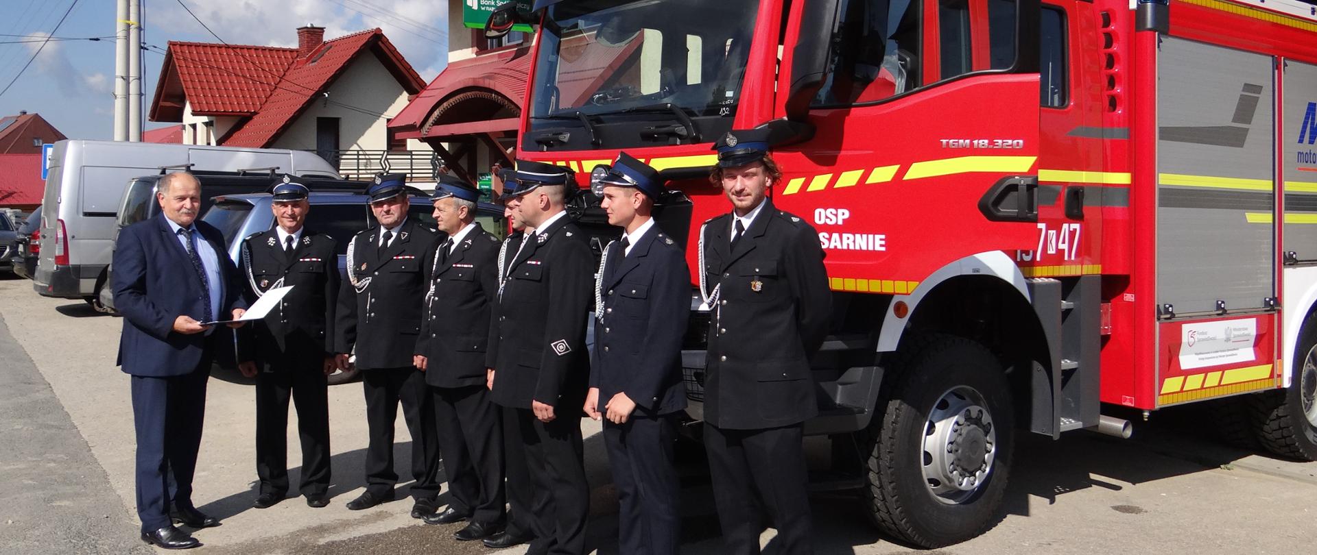 Zdjęcie przedstawiające tle remizy strażackiej Wójta Gminy przekazującego samochód bojowy dla strażaków z Podsarnia.
