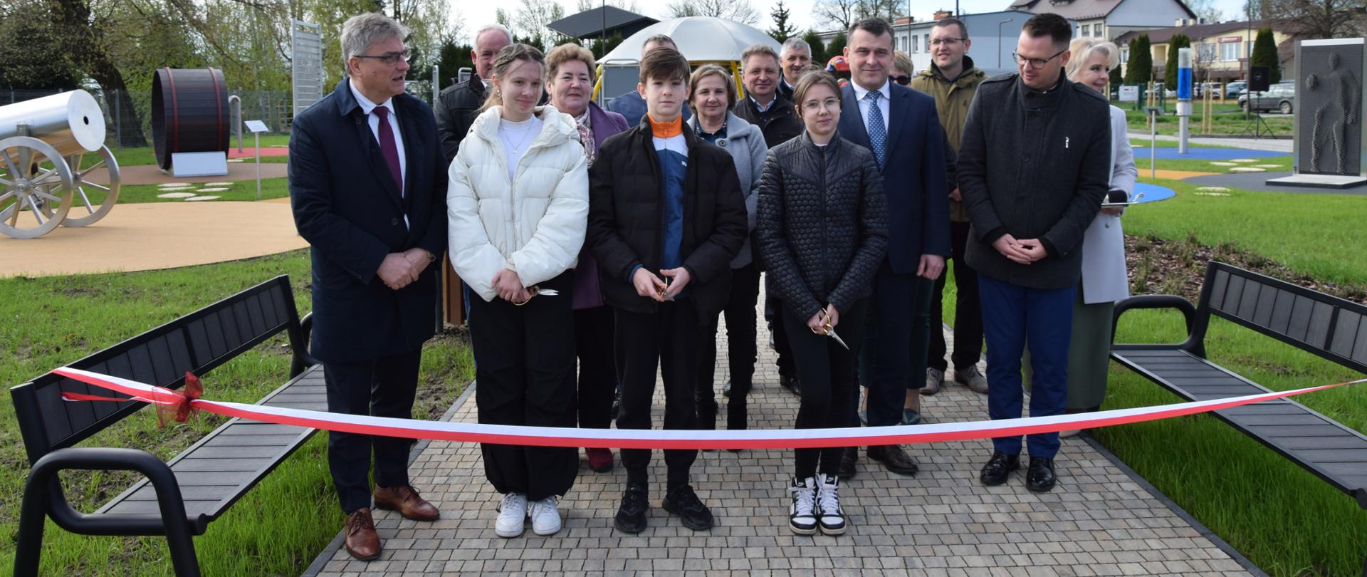 Zdjęcie przedstawia osoby w wieku od 14 do 65 lat , dzieci, kobiety i mężczyzn stojących za wstęgą rozwieszoną pomiędzy dwoma ławkami.