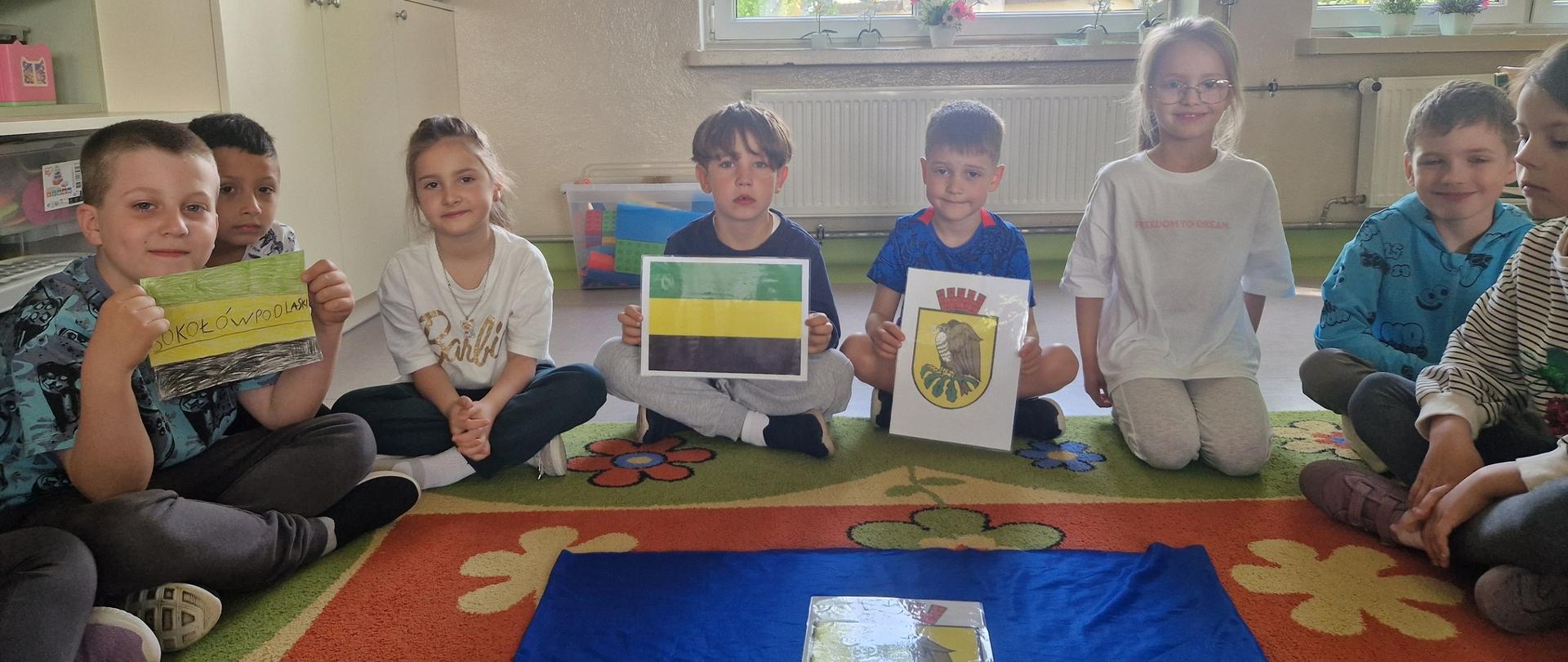8 dzieci siedzi na dywanie z flagą i herbem miasta