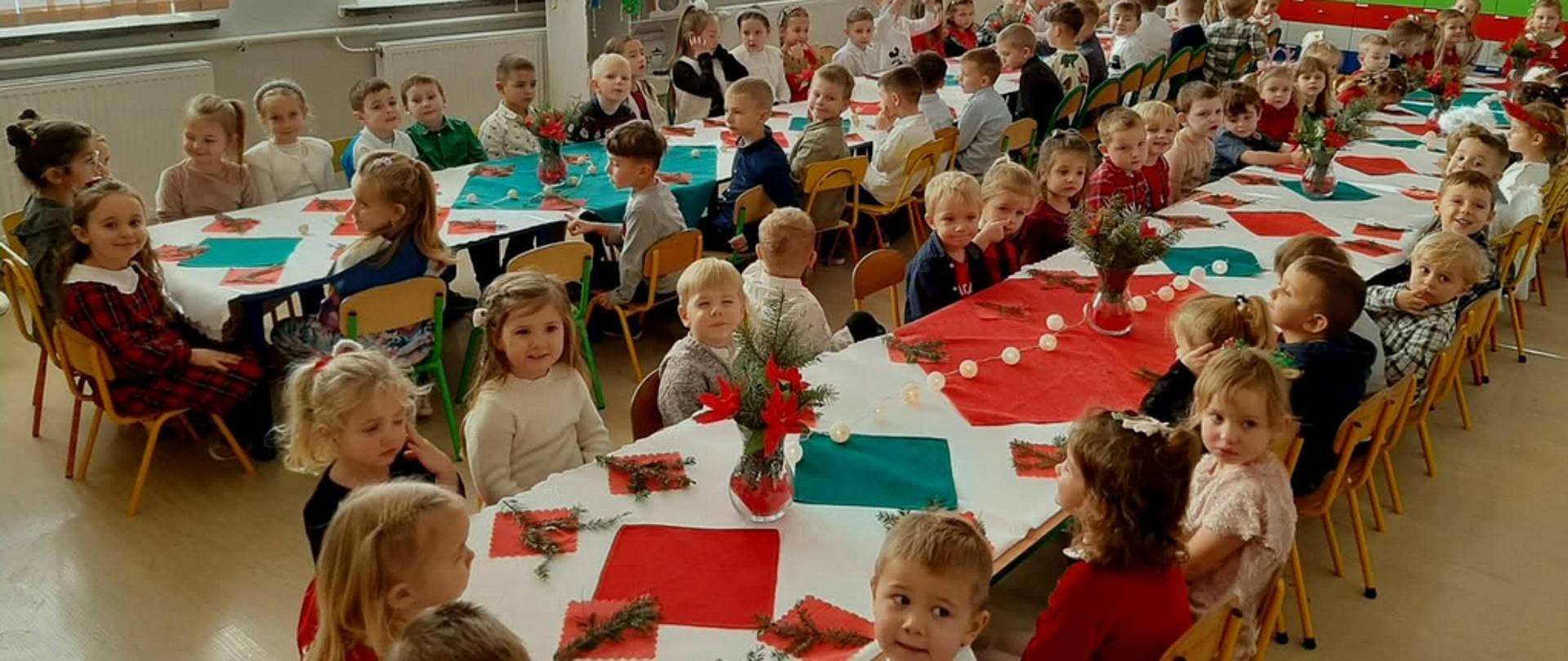 Duża grupa dzieci siedzących przy długich stołach udekorowanych białymi i czerwonymi obrusami oraz stroikami z gałązek iglastych. Uczestnicy spotkania są odświętnie ubrani i wspólnie świętują w jasnej sali, w której znajduje się ubrana choinka oraz dekoracje ścienne z motywem kosmosu. W tle widoczne są osoby dorosłe przygotowujące posiłek lub doglądające przebiegu uroczystości.