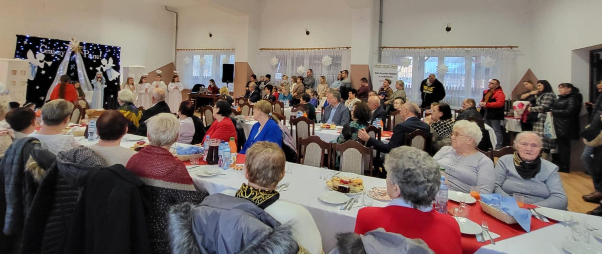 Zdjęcie przedstawia uroczyste spotkanie w udekorowanej sali. Przy stołach siedzą elegancko ubrani goście, a w tle stoją dorośli i dzieci. Atmosfera jest oficjalna i rodzinna.