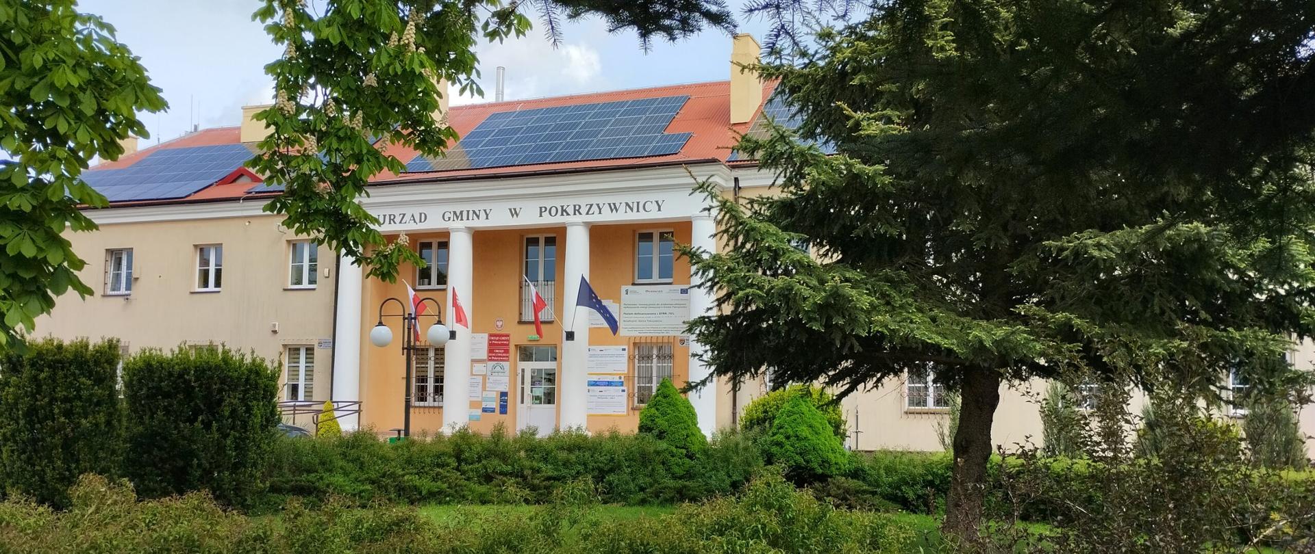 Urząd Gminy Pokrzywnica to budynek z beżową fasadą i dwoma dużymi kolumnami przy wejściu. Nad drzwiami znajdują się tablice informacyjne, a obok powiewają trzy flagi: biało-czerwona, niebieska oraz z herbem. Na dachu zamontowane są panele słoneczne. Budynek otoczony jest zielenią – drzewami i krzewami.