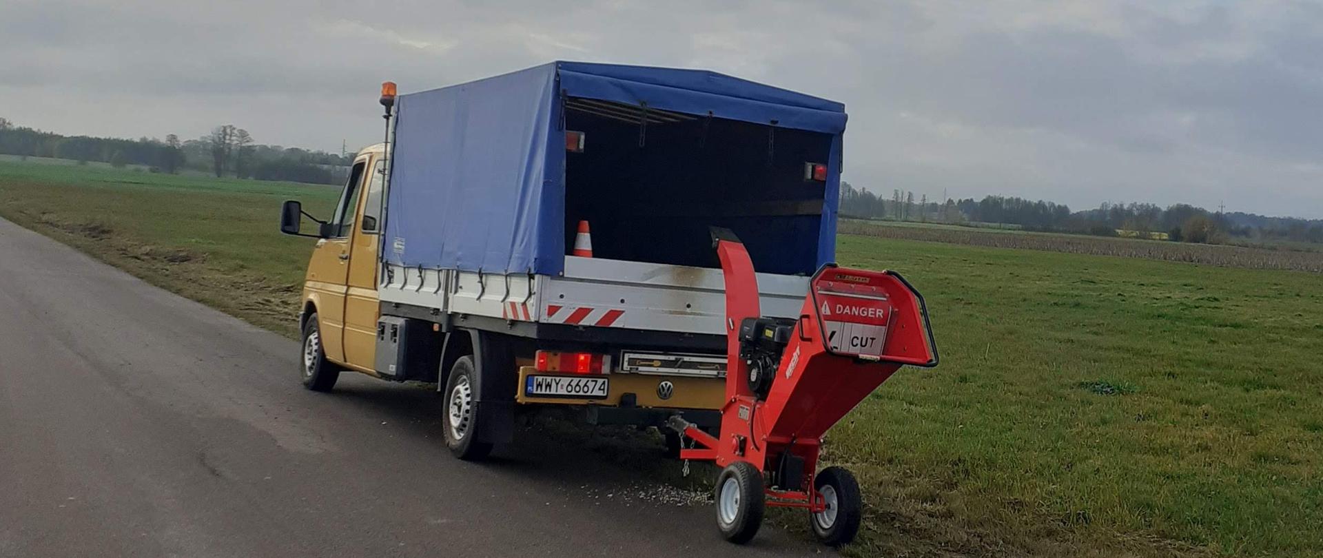 Zdjęcie przedstawia siedmioosobowy samochód dostawczy VW Doca wraz z zaczepionym do niego rębakiem. Samochód jest koloru żółtego z zabudową skrzyniową oraz plandeką w kolorze ciemnoniebieskim. Do samochodu zaczepiony jest czerwony rębak, który posiada silnik spalinowy oraz dwa koła. W tle widać łąki, pola uprawne drzewa oraz ulicę.