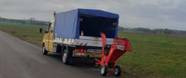 Zdjęcie przedstawia siedmioosobowy samochód dostawczy VW Doca wraz z zaczepionym do niego rębakiem. Samochód jest koloru żółtego z zabudową skrzyniową oraz plandeką w kolorze ciemnoniebieskim. Do samochodu zaczepiony jest czerwony rębak, który posiada silnik spalinowy oraz dwa koła. W tle widać łąki, pola uprawne drzewa oraz ulicę.