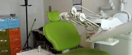 Zielony fotel stomatologiczny . Obok niego stoi stolik na jednej nóżce oraz przy nim wiszą narzędzia dentystyczne