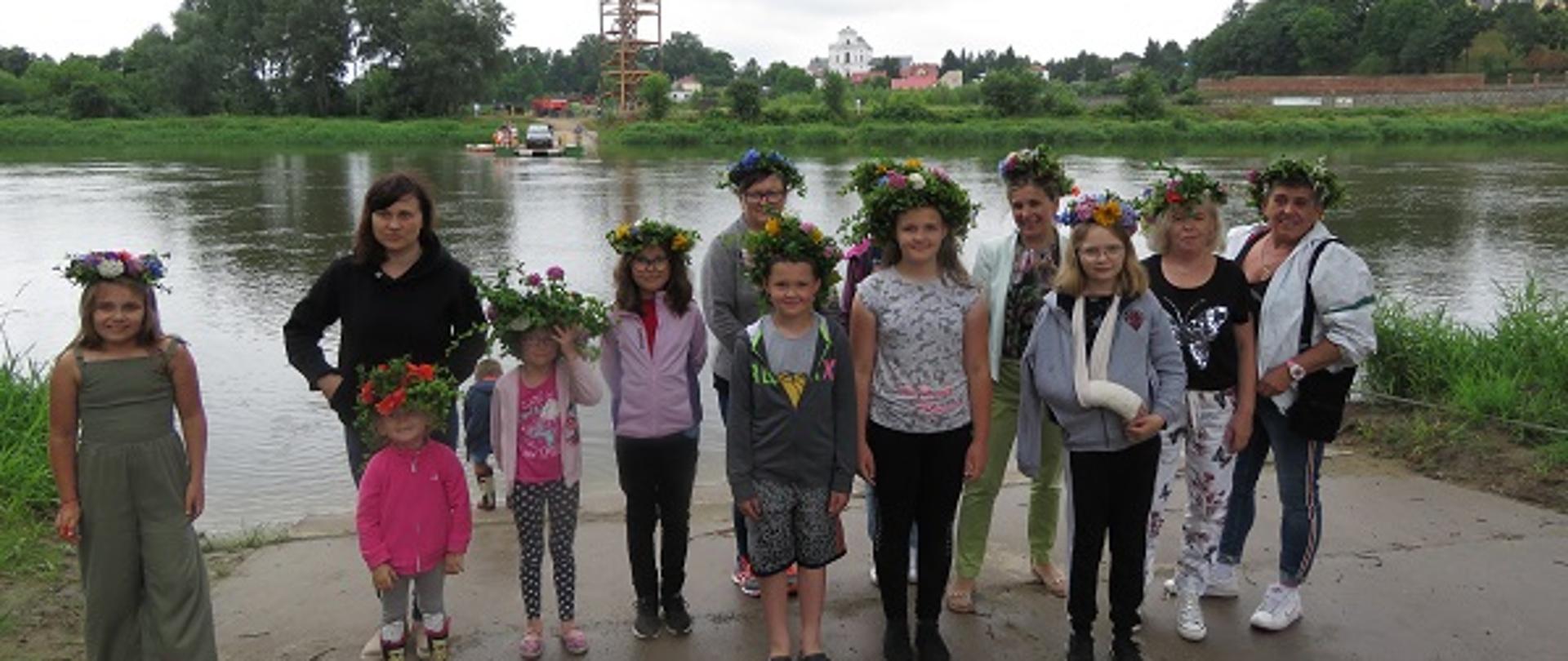 W tle rzeka. Na pierwszym planie grupa kobiet i dzieci z wiankami z kolorowych kwiatów na głowach.
