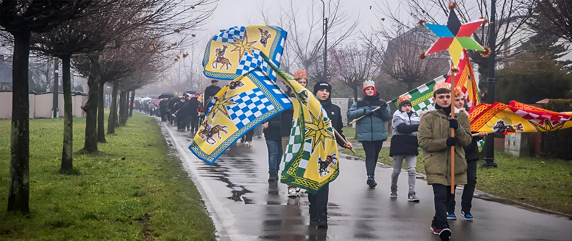 Młodzież idąca po drodze z kolorowymi flagami. Obok zielony trawnik i drzewa bez liści.