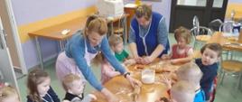 Na zdjęciu dzieci siedzą przy stolikach i przygotowują ciastka do pieczenia, pomagają im dwie panie.