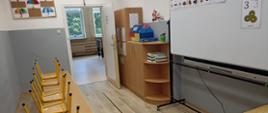 Mała sala lekcyjna z jasnymi panelami na podłodze i popielatymi ścianami, po lewej stronie 3 ławki uczniowskie z krzesłami, po prawej stronie ruchoma biała tablica na kółkach, za nią szafka z pomocami dydaktycznymi, na wprost otwarte drzwi na korytarz