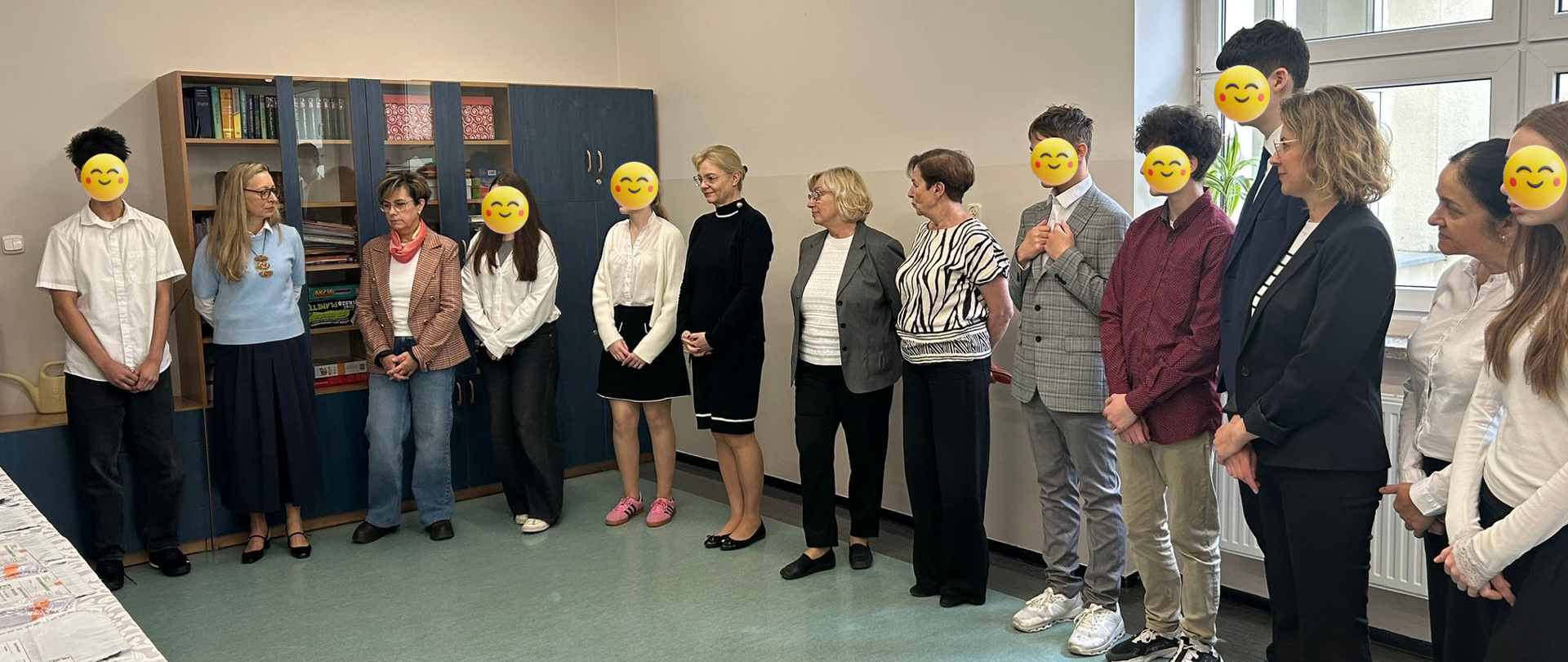 Po lewej stronie widać stół przykryty obrusem. Na nim leżą egzaminy. Po prawej stronie w półkolu stoją panie i uczniowie, którzy mają twarze zasłonięte uśmiechniętymi minkami. 