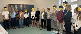 Po lewej stronie widać stół przykryty obrusem. Na nim leżą egzaminy. Po prawej stronie w półkolu stoją panie i uczniowie, którzy mają twarze zasłonięte uśmiechniętymi minkami. 