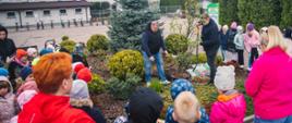 Dwoje mężczyzn stoi przy drzewku. Jeden trzyma w górze szpadel. W koło nich stoi grupa dzieci i 2 kobiety