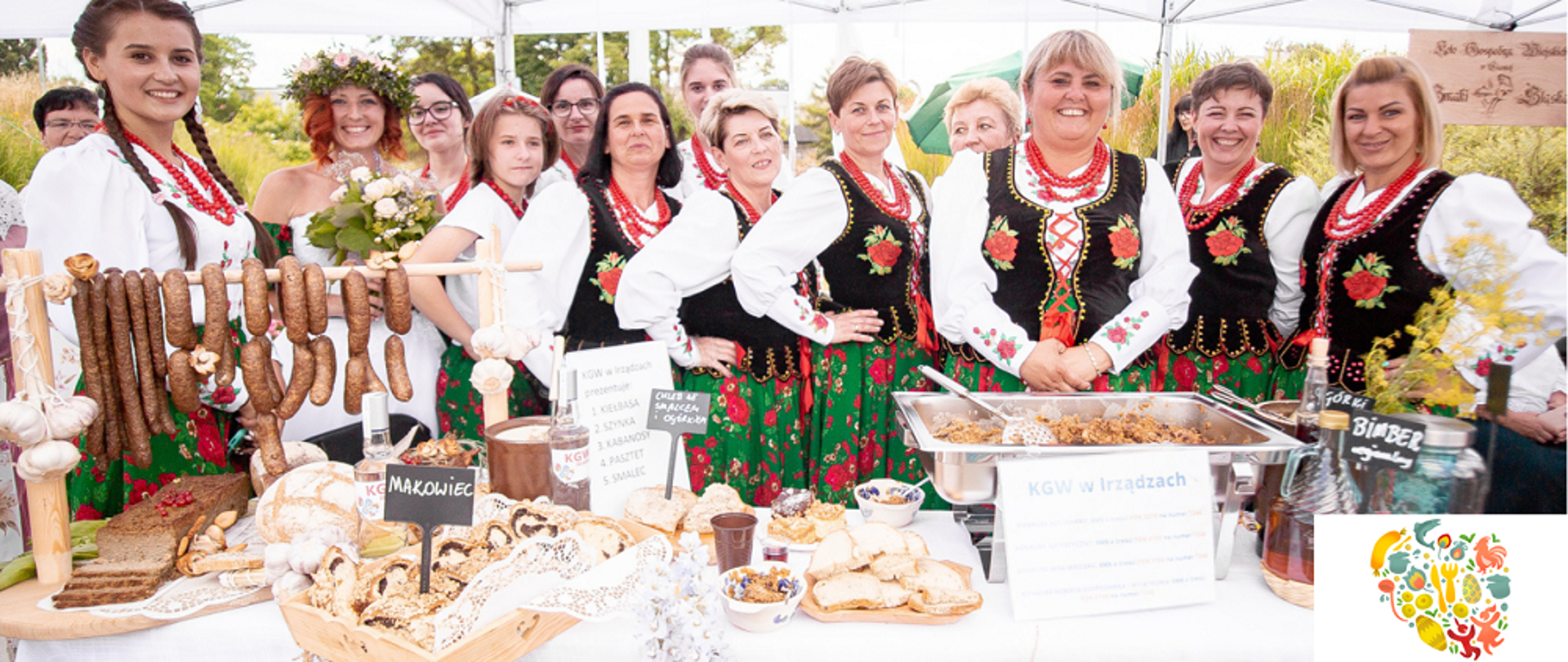 kobiety przed stołem zastawionym różnymi wyborami (kiełbasa, smalec). Panie ubrane w czarne gorsety, białe bluzki i zielone spódnice