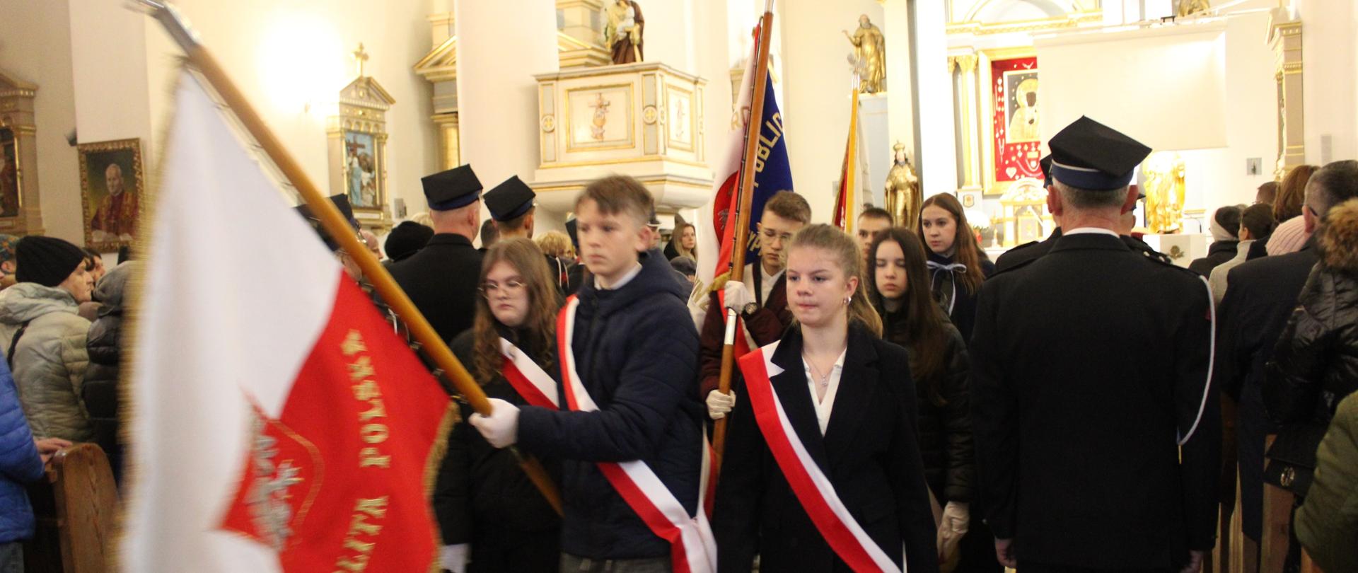 Młodzież wychodzi z kościoła. Chłopcy niosą w rękach uniesione sztandary. Przepasani są biało czerwonymi wstążkami
