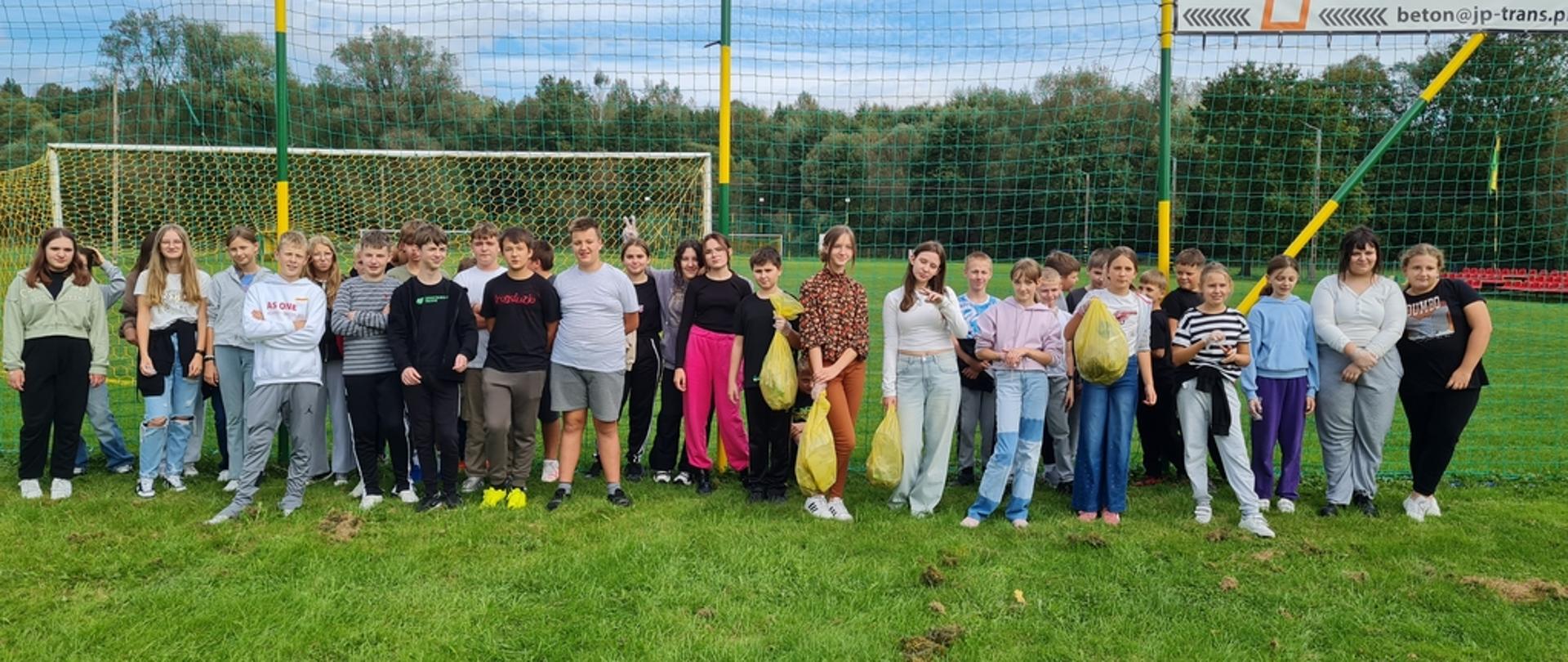 Duża grupa dzieci z klasy piątej i siódmej pozuje do zdjęcia przed boiskiem sportowym. Niektórzy trzymają w rękach żółte worki ze śmieciami.