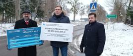 Minister Maciej Małecki wraz ze Starostą Krzysztofem Bieńkowskim i Członkiem Zarządu Powiatu Przasnyskiego Pawłem Mostowym prezentują środki przyznane w ramach programu Polski Ład. Zdjęcie wykonano w pobliżu Szkoły Podstawowej w Bartnikach przy drodze powiatowej prowadzącej w kierunki Jednorożca i Baranowa. Starosta i Minister trzymają tablice informujące o wysokości przyznanych środków finansowych. Od lewej: Starosta, Minister, Członek Zarządu Powiatu Przasnyskiego.