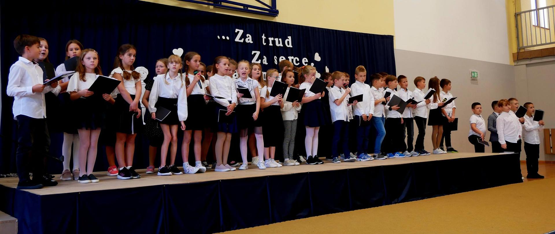 Trzecioklasiści na scenie śpiewają piosenkę dla nauczycieli i pracowników szkoły.