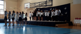 Na scenie stoją elegancko ubrani uczniowie z klasy 6 chłopcy i dziewczęta. Przed nimi uczennica śpiewa do mikrofonu. Przed sceną na 7 uczennic stoi jedna za drugą w rękach mają szarfy, które powiewaja w powetrzu.