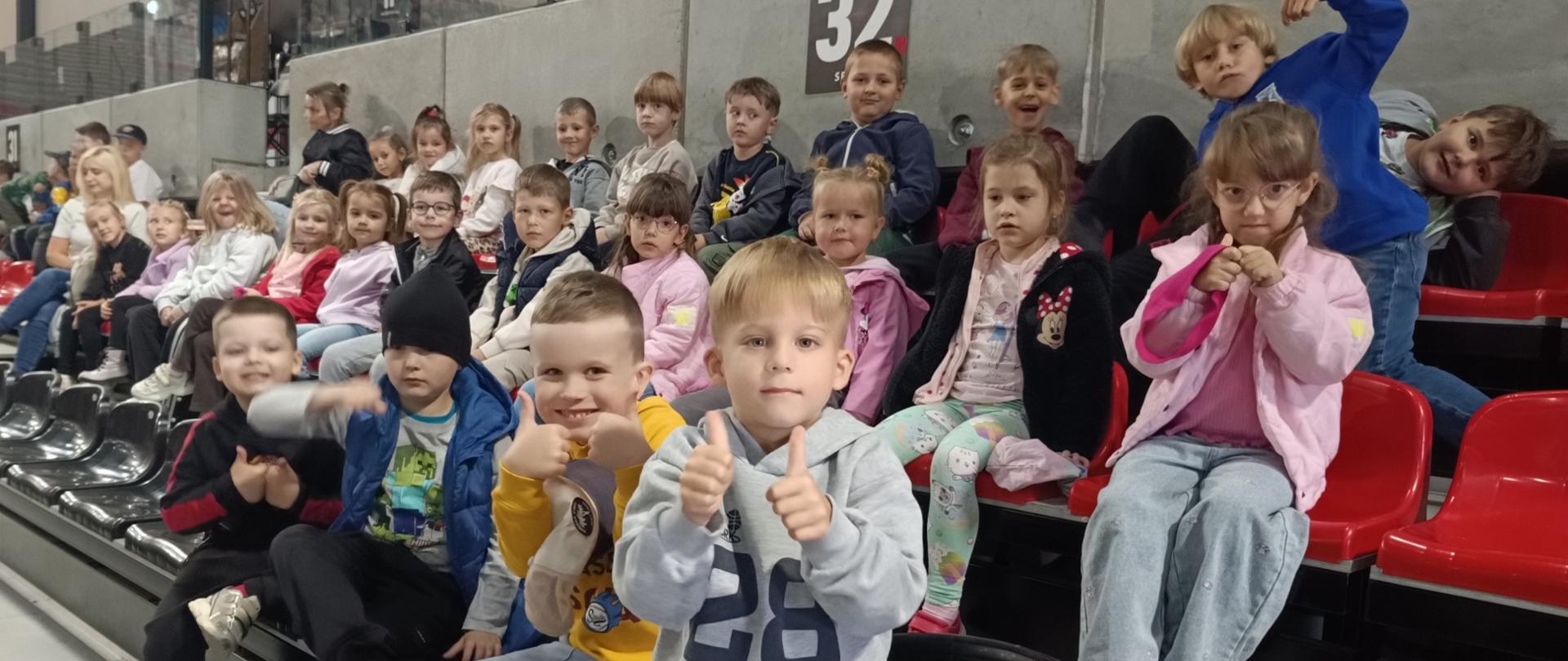 Grupa dzieci w wieku przedszkolnym siedzi na trybunach w hali sportowej, z których część uśmiecha się i pokazuje kciuki do góry.