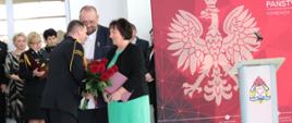 Fotografia wykonana w garażu PSP w Sokołowie Podlaskim podczas uroczystego pożegnania komendanta. Na zdjęciu moment podziękowań i wręczenia kwiatów przez przewodniczącego Rady Powiatu Sokołowskiego oraz starostę, komendantowi odchodzącemu na emeryturę. Po lewej stronie widać część zaproszonych gości, natomiast po prawej mównicę i czerwone tło z wizerunkiem orła białego.