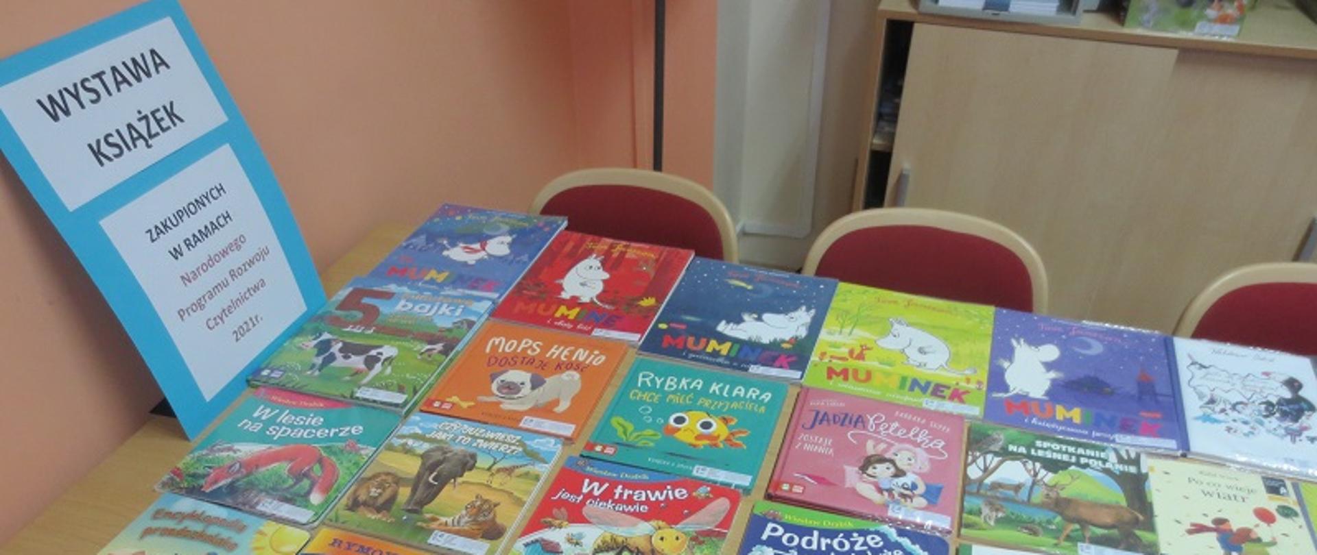 Zdjęcie przedstawia książki dla dzieci ułożone na ławce. Z lewej strony plakat z napisem wystawa książek