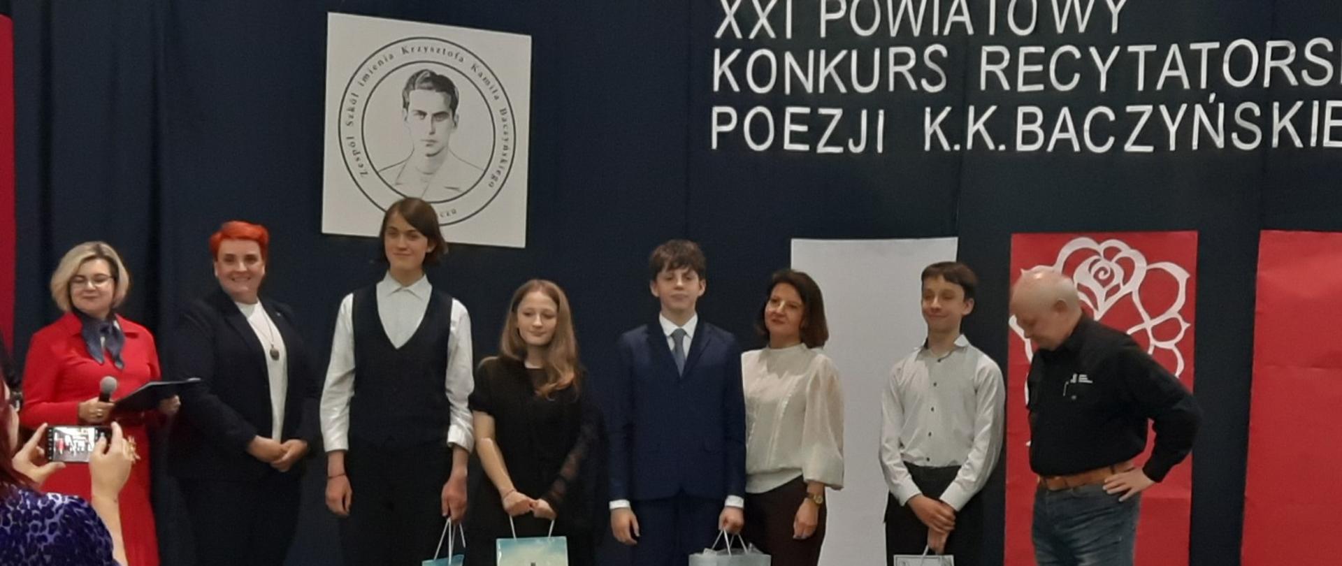 Grupa młodzieży stojąca obok siebie. W rękach trzymają o jednej papierowej torbie. Obok nich stoi mężczyzna i 2 kobiety. Za nimi napis XXI Powiatowy konkurs recytatorski poezji KK Baczyńskiego