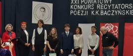Grupa młodzieży stojąca obok siebie. W rękach trzymają o jednej papierowej torbie. Obok nich stoi mężczyzna i 2 kobiety. Za nimi napis XXI Powiatowy konkurs recytatorski poezji KK Baczyńskiego