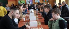 Zdjęcie przedstawia grupę graczy, którzy siedząc przy stolikach na przeciw siebie rozgrywają partie turnieju szachowego