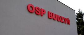 ściana szczytowa budynku w kolorze szarym, z podświetlanym, czerwonym napisem "OSP Budzyń"