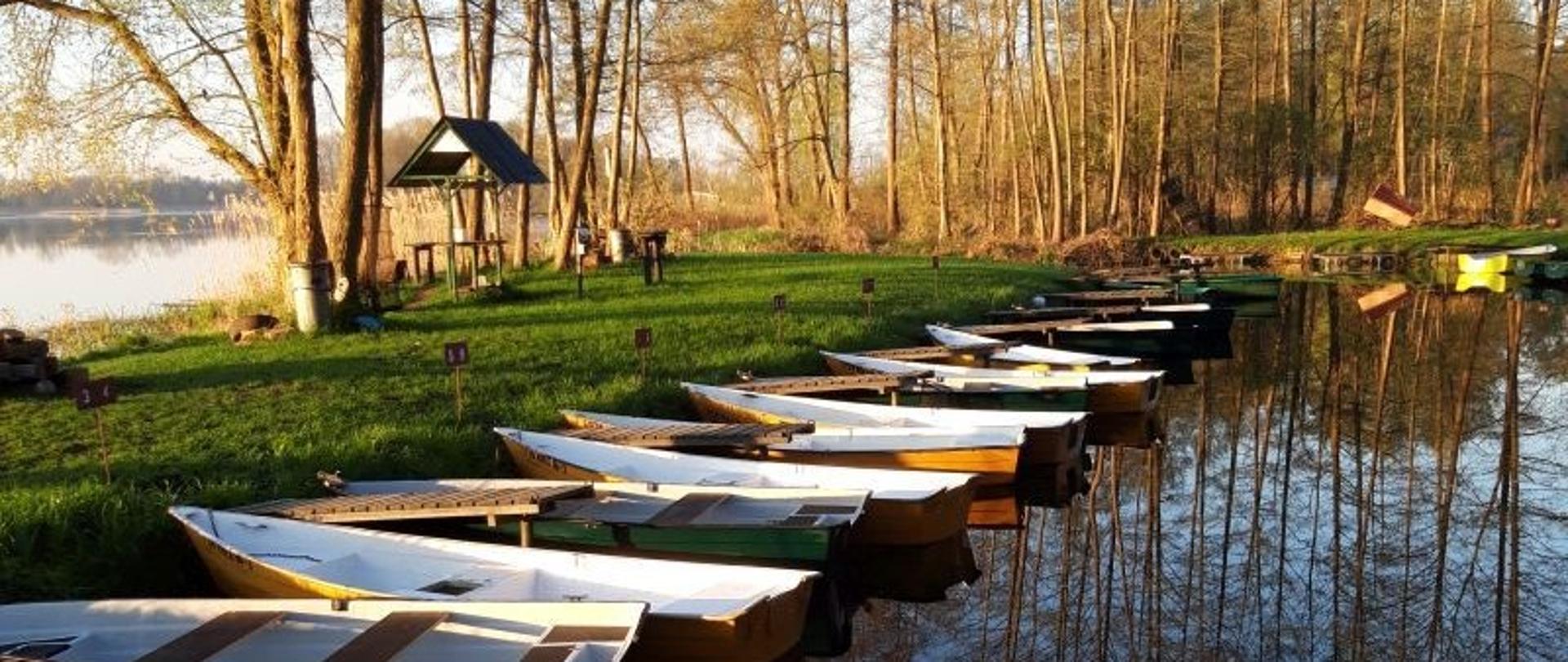 Zdjęcie przedstawia spokojną scenę nad jeziorem. Wzdłuż brzegu w wodzie zacumowane są małe łodzie, ustawione równolegle do siebie, odbijające się w tafli jeziora. W tle widoczna jest trawiasta polana z wysokimi drzewami oraz drewnianym schronieniem lub altaną. Całość oświetlona jest delikatnym światłem słonecznym, nadającym zdjęciu cichy i spokojny charakter.
