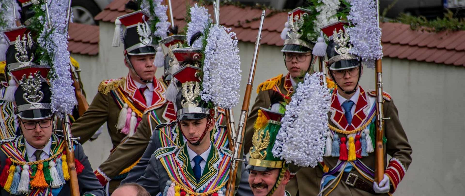 mężczyźni ubrani w ciemnozielone garnitury z kolorowymi pomponami i lampasami robią pokazy musztrowe. na głowie mają czapki z dużym białym kwiatem